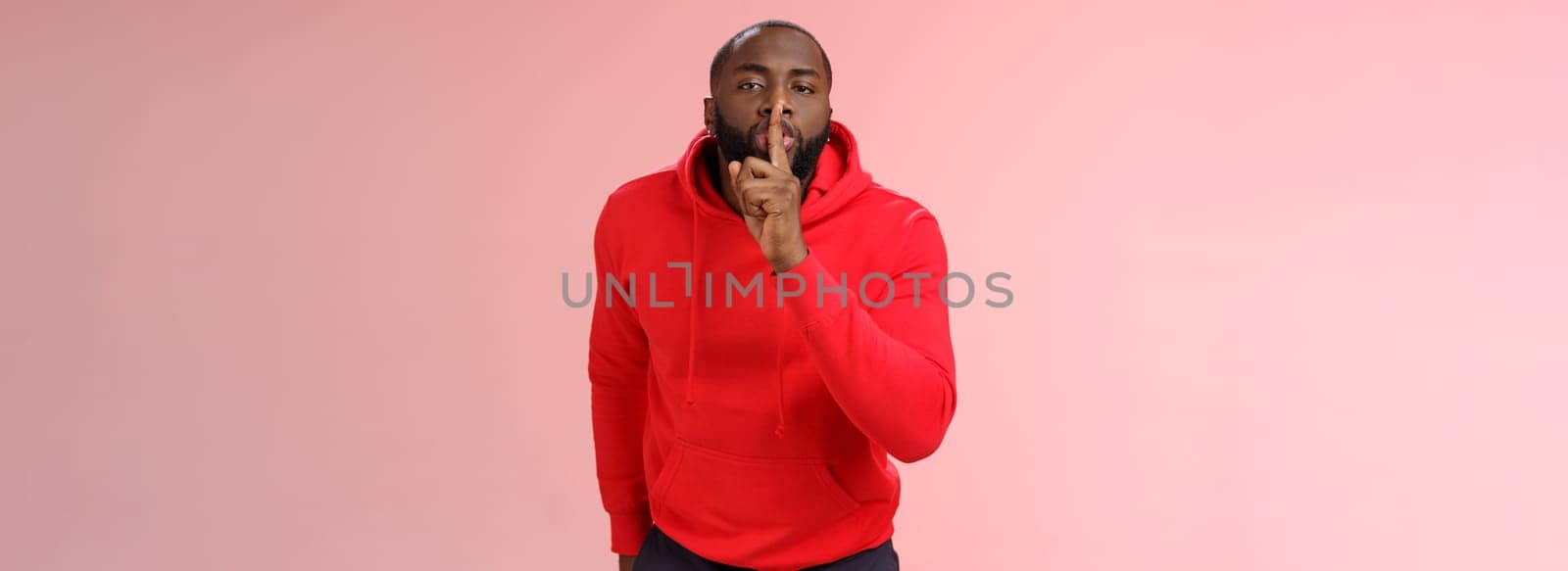 Shhh it taboo. Portrait sensual funny cute african-american bearded man in red hoodie bending camera show shush gesture index finger on lips telling secret asking keep quiet not tell anyone mystery.