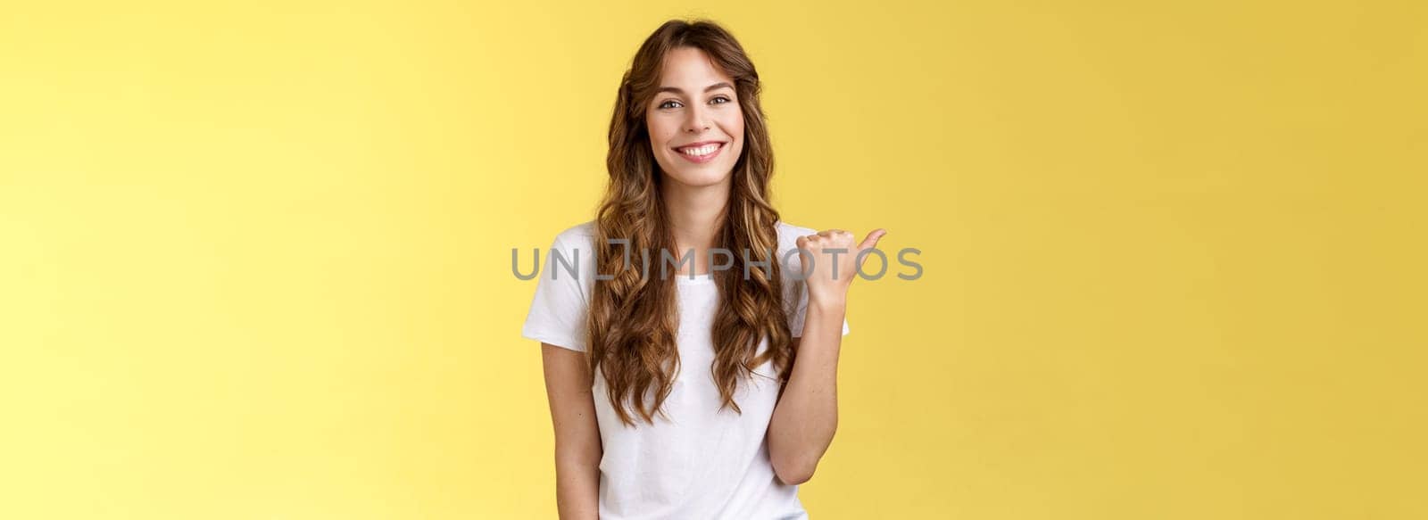 Visit see yourself. Cheerful charismatic good-looking outgoing girl long curly haircut showing place do good hairstyle smiling happily delighted pointing thumb left introduce promo yellow background.