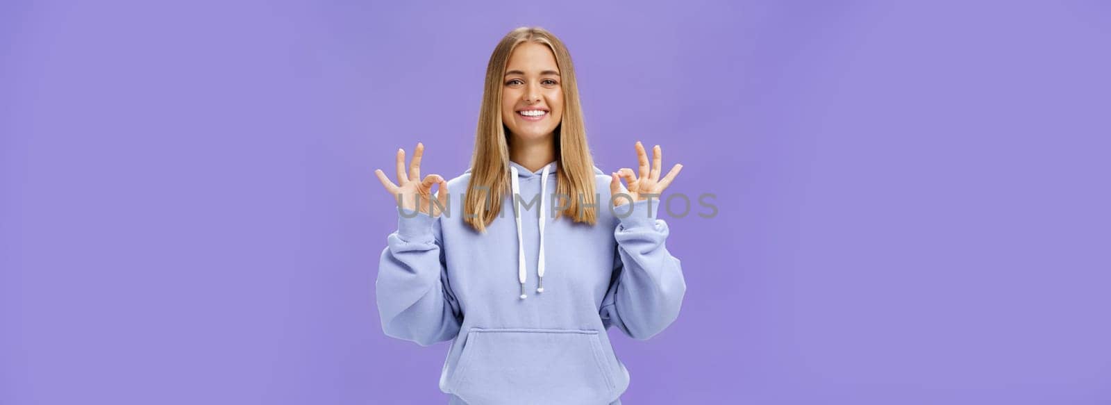 Girl got everything under control. Delighted happy charming woman with fair hair and tanned skin without makeup showing okay gesture smiling assured and pleased posing in hoodie over purple wall.