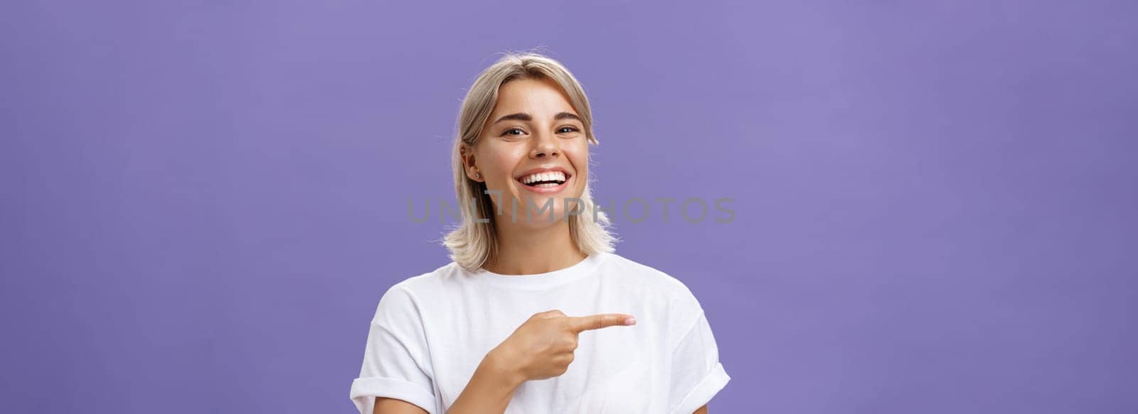 Close-up shot of amused happy and entertained good-looking sociable woman with fair hair and beautiful smile grinning while pointing left with index finger showing awesome copy space over purple wall by Benzoix