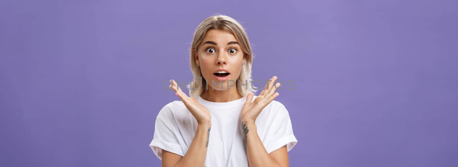 Close-up shot of charmed and impressed good-looking caucasian girlfriend with blond hair and tattoos on arms raising hands in amazed and excited gesture opening mouth and gazing mesmerized at camera by Benzoix
