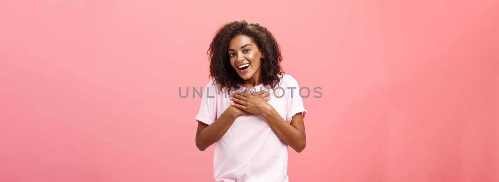 Portrait of charming delighted african american woman with curly haircut holding palms on heart pleased and grateful thanking friend for help smiling happily and thankful at camera over pink wall by Benzoix