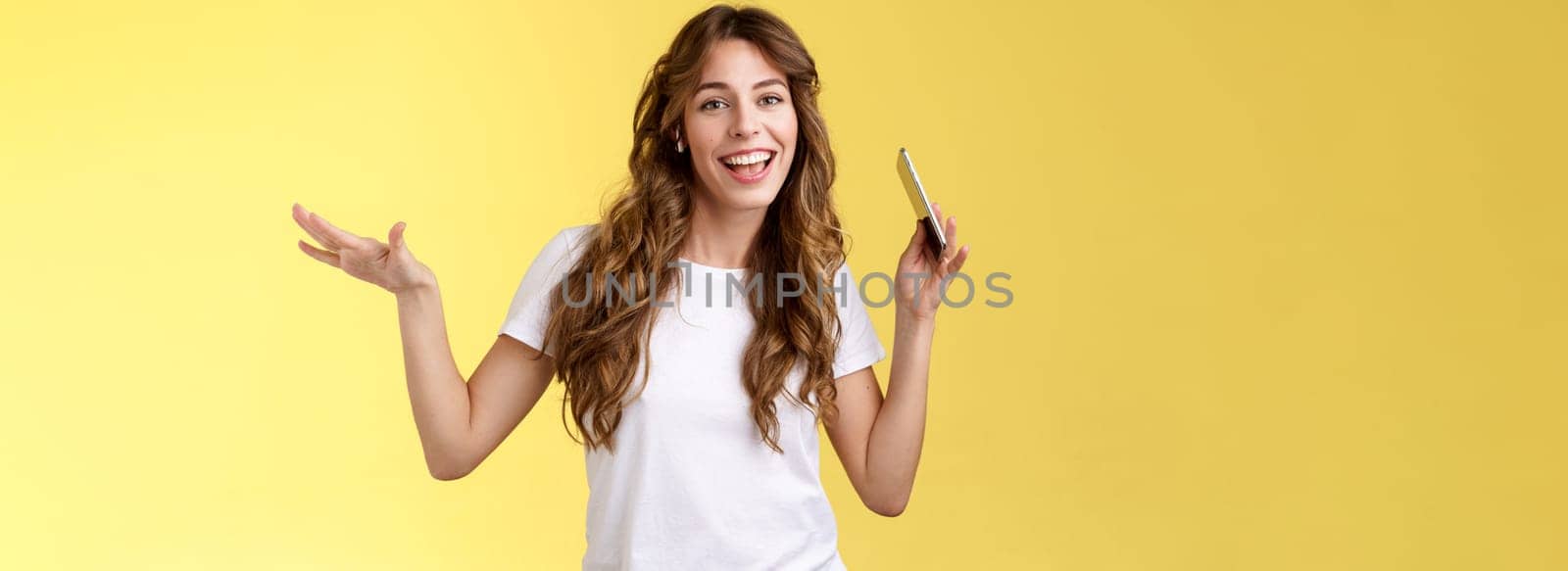 Sassy cheerful carefree attractive modern girl curly hairstyle having fun lively upbeat mood moving body rhythm shake hands hold smartphone lip sync wear wireless earbuds listen music dancing. Lifestyle.