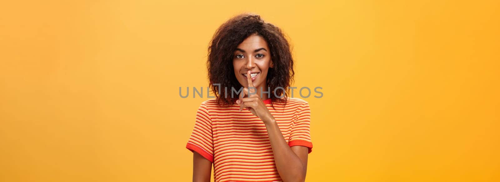 Not say anyone till I give permission. Happy pleased and mysterious attractive dark-skinned female with curly hairstyle telling beauty secret to friend making shh sound with index finger over mouth.