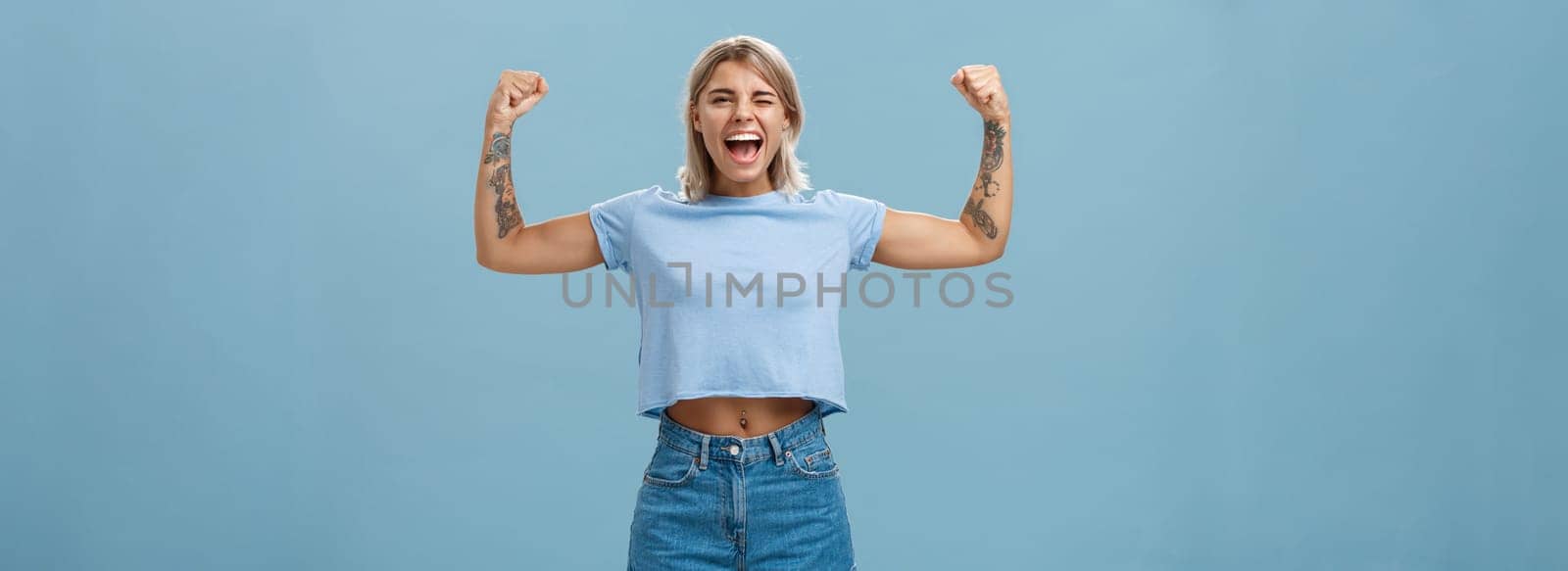 Strong women stand up and fight for rights. Portrait of happy entertained and cool young female athletic blonde with tattoos winking and smiling showing muscles and biceps over blue wall.