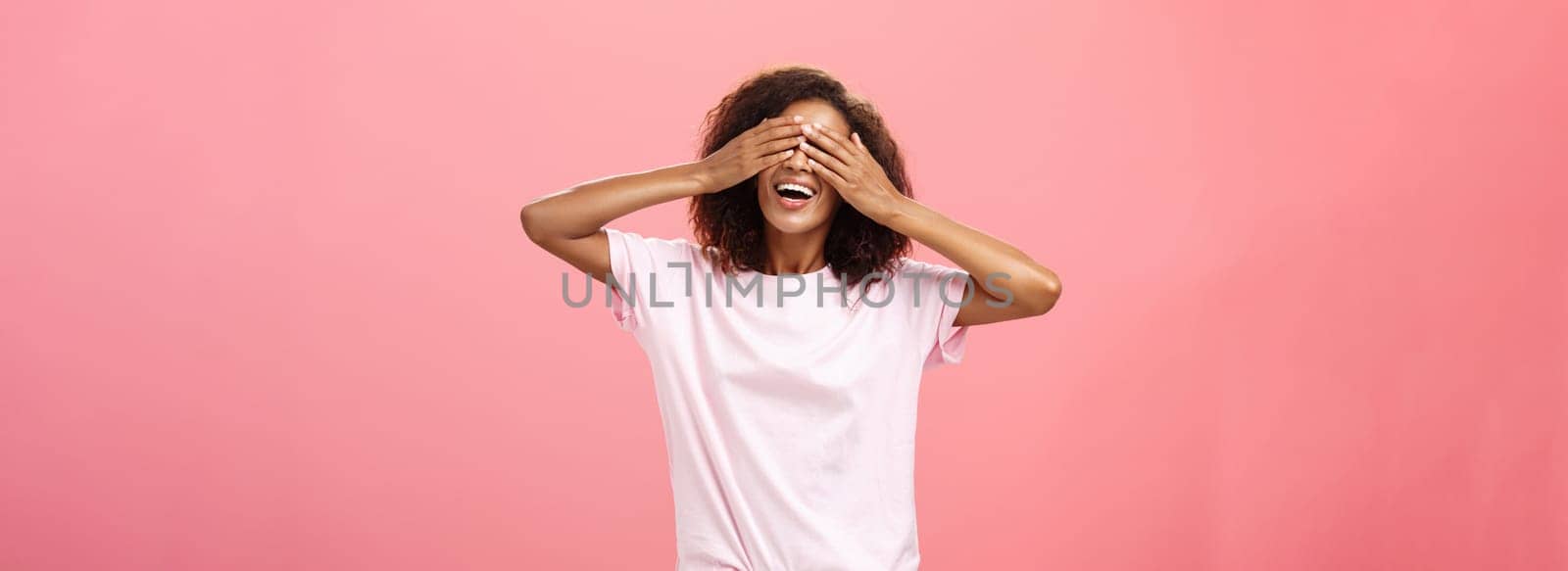 Portrait of charming joyful dark-skinned playful woman with curly hair in t-shirt closing eyes and counting ten with broad happy smile playing hide n seek or waiting for surprise over pink background by Benzoix