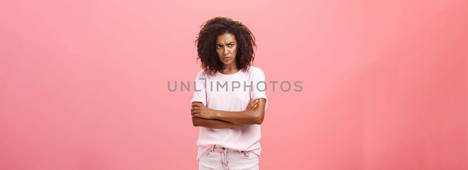 Portrait of offended gloomy sad african american female friend crossing arms on chest in protection gesture frowning looking from under forehead with insult looking envious and angry over pink wall. Lifestyle.