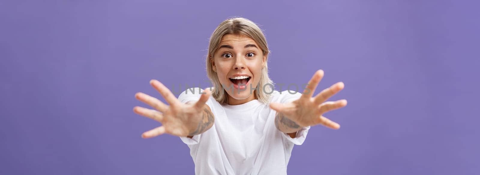 Lifestyle. Waist-up shot of amused and excited attractive stylish young woman in white t-shirt pulling hands at camera with desire smiling thrilled and happy wanting hug or take something over purple background.