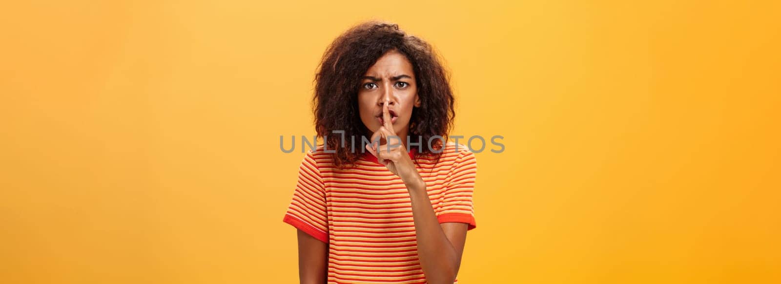 Shh do not disturb. Portrait of serious-looking bossy and displeased cute african american girl with afro hairstyle frowning shushing at camera with index finger over mouth forbidding speak loud by Benzoix