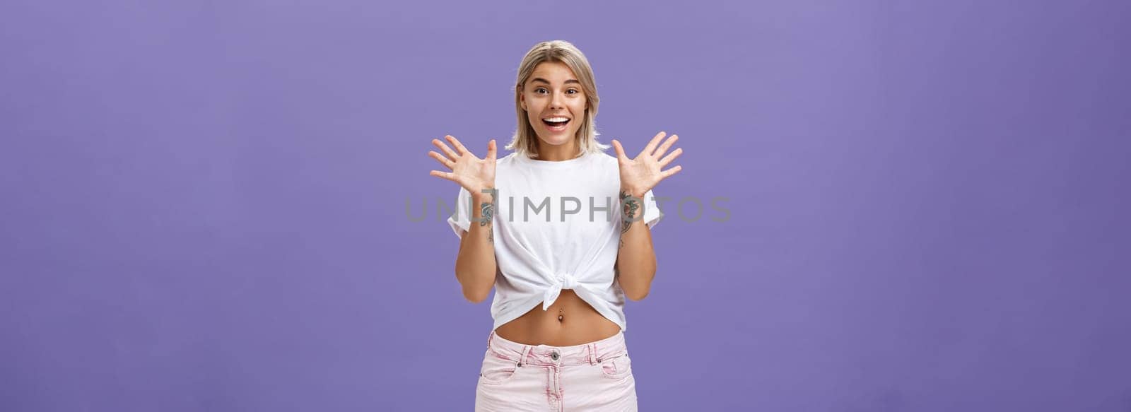 Lifestyle. Indoor shot of delighted and amused attractive stylish urban female with blond hair in stylish white t-shirt and summer shorts raising palms from surprise and amazement standing over purple background.