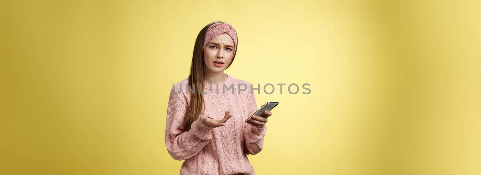 Puzzled confused upset questioned young cute girl in headband knitted sweater frowning clueless raising hand in dismay holding smartphone, cannot understand what happened cellphone over yellow wall by Benzoix