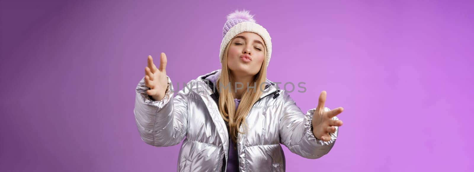 Romantic charming caucasian blond woman in cute hat silver stylish jacket pulling arms forward wanna embrace tight cuddle boyfriend folding lips close eyes give kiss, standing purple background by Benzoix
