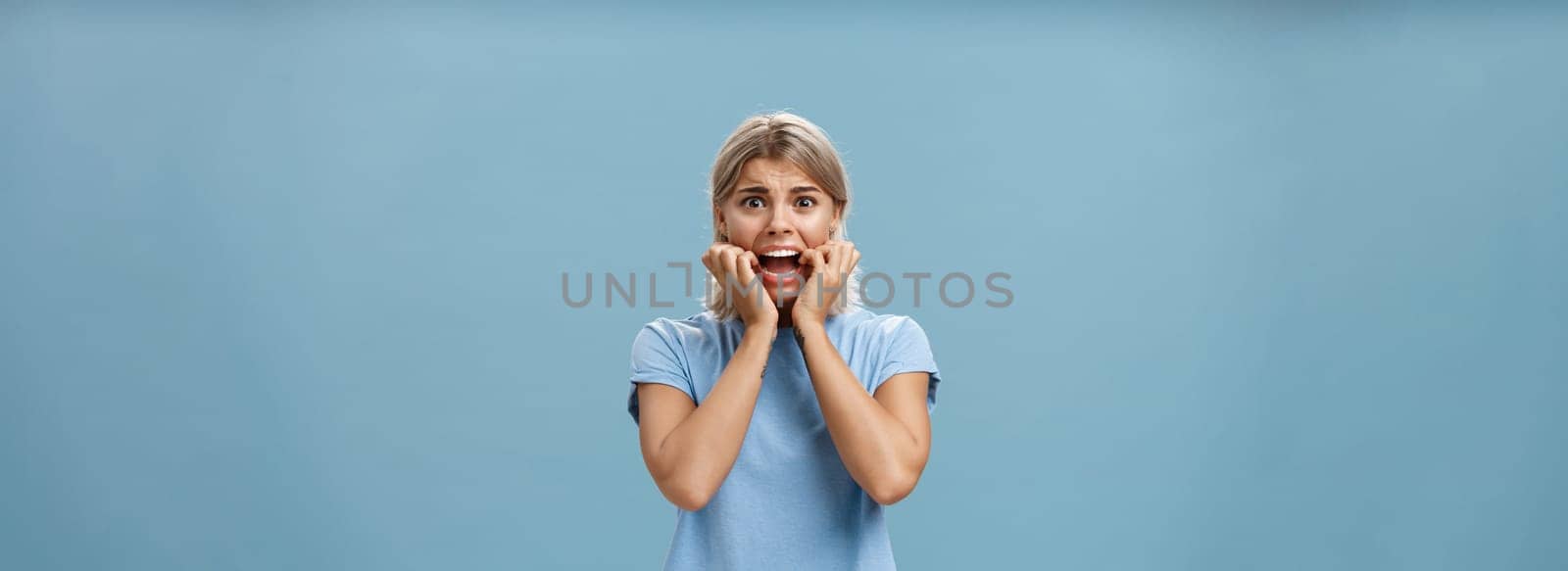 Indoor shot of shocked troubled girlfriend hearing mom returned home being with boyfriend biting fingernails being in helpless perplexed situation feeling anxious and scared over blue background by Benzoix