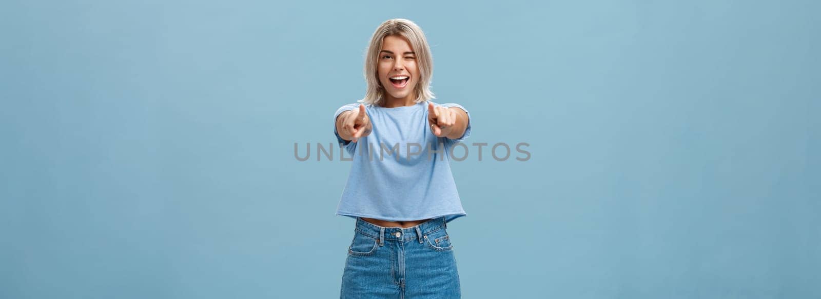 Happy enthusiastic stylish blonde female with tattoos pointing at camera as if picking or making hint smiling joyfully and winking from amazement and happiness posing over blue background by Benzoix