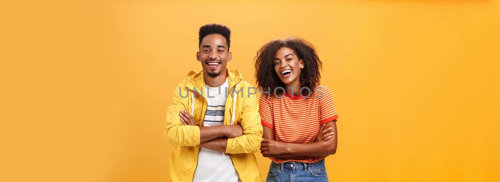 Two african american man and woman being best friends laughing out loud watching funny movie in cinema all dressed up in stylish outfit. standing with hands crossed on chest and amused expression.