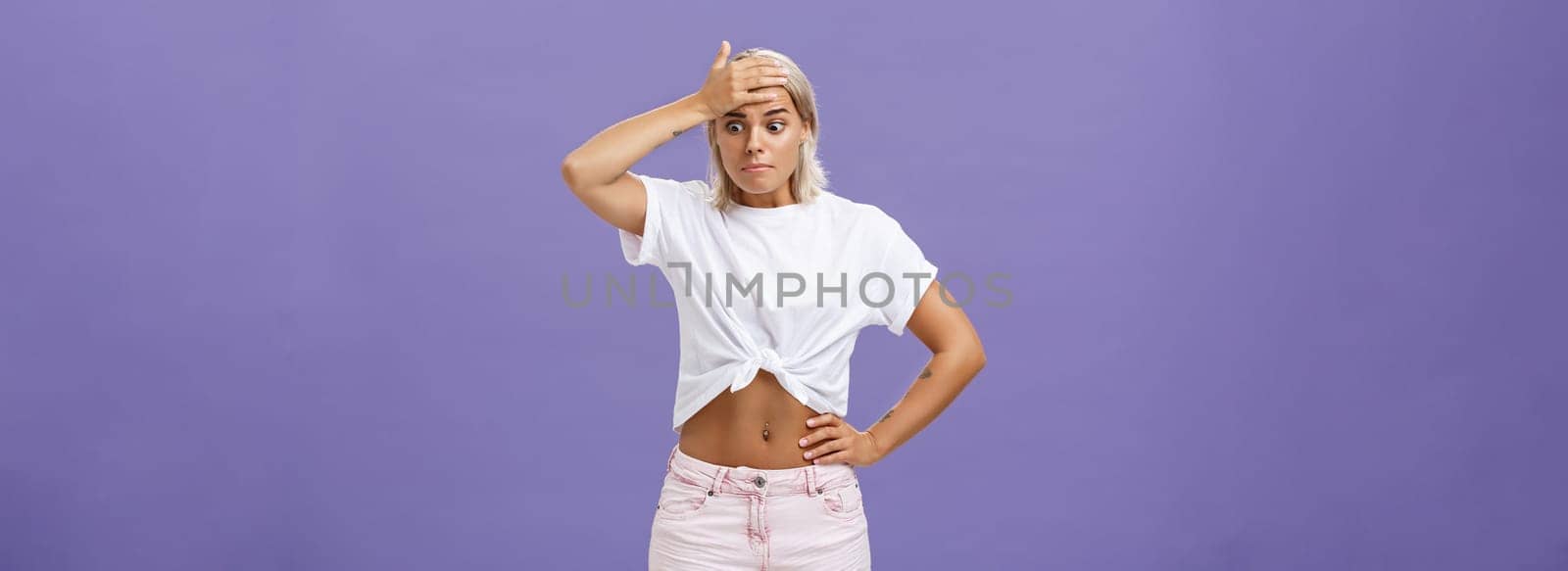 Woman got in serious bad sitation feeling concerned and nervous starting panic holding hand on forehead pursing lips staring down with shocked, perplexed expression being in trouble over purple wall.