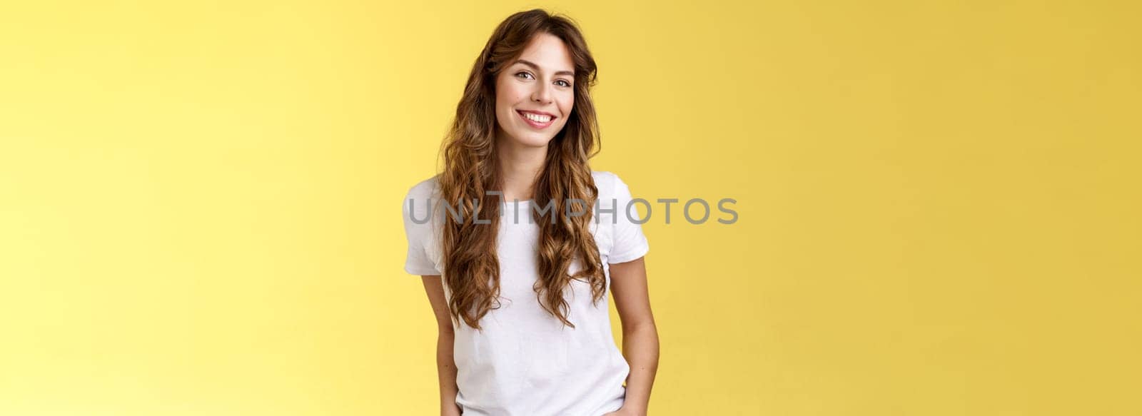 Attractive carefree confident friendly-looking charming feminine ubran woman long curly hair smiling toothy pleased tilt head intrigued hold hands pockets self-assured pose stand yellow background. Lifestyle.