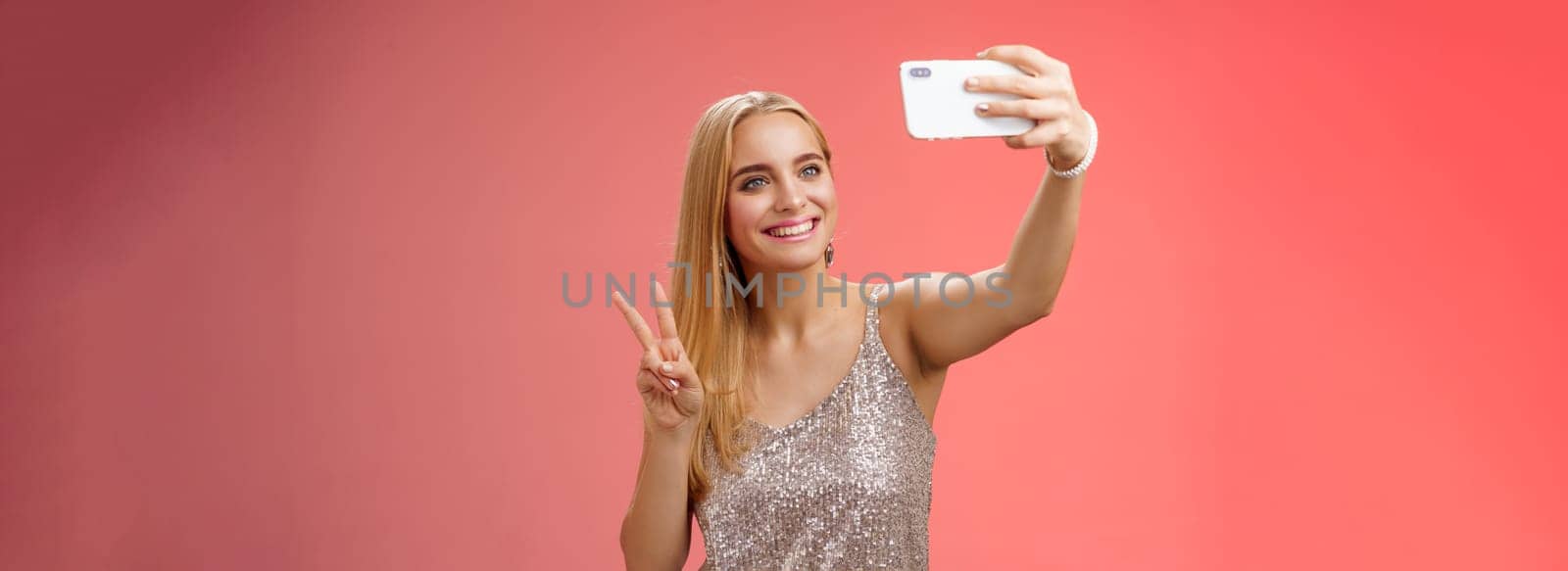 Attractive feminine tender young blond girl 25s in silver stylish dress taking selfie extend arm up show peace gesture smartphone display record video greeting internet fans, standing red background.