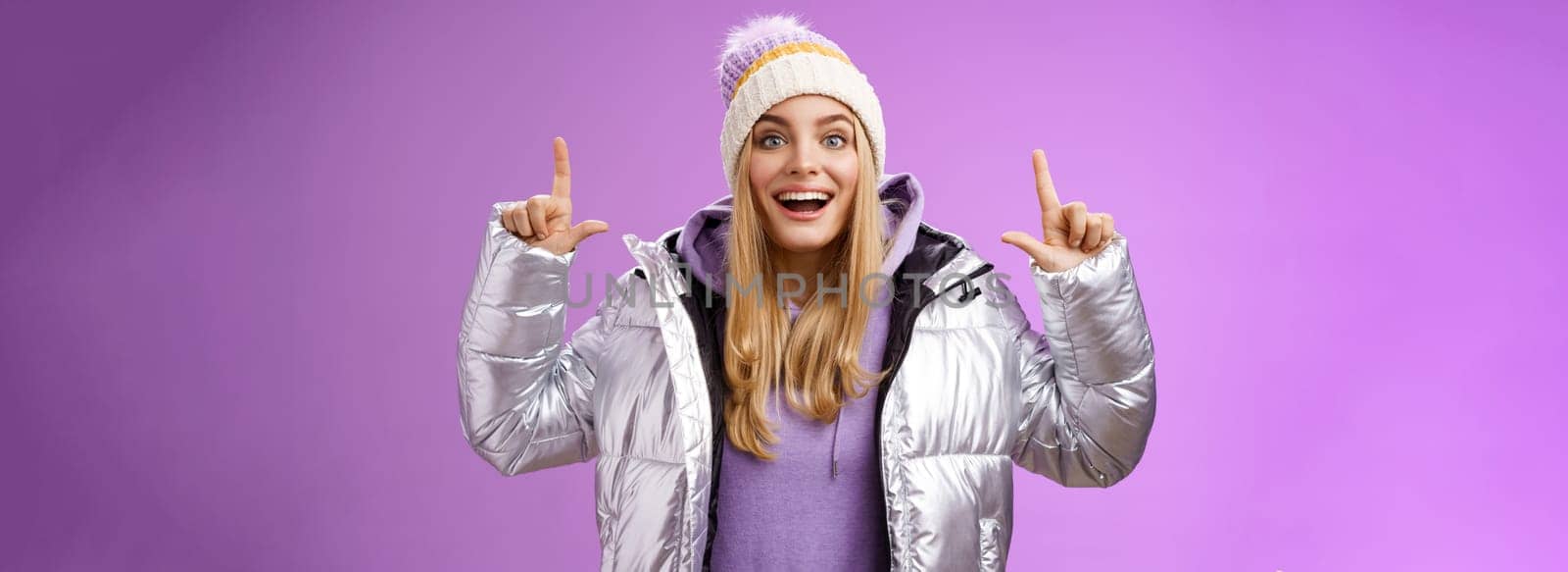Lifestyle. Excited carefree cheerful fair-haired european girl in silver jacket winter hat raising hands pointing up have excellent idea smiling broadly speaking passionately standing purple background.