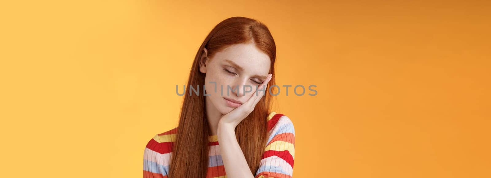 Tired cute redhead female student exhausted feel sleepy fall asleep standing leaning face palm close eyes working part-time night shift, daydreaming lacking energy wanna sleep bed orange background.