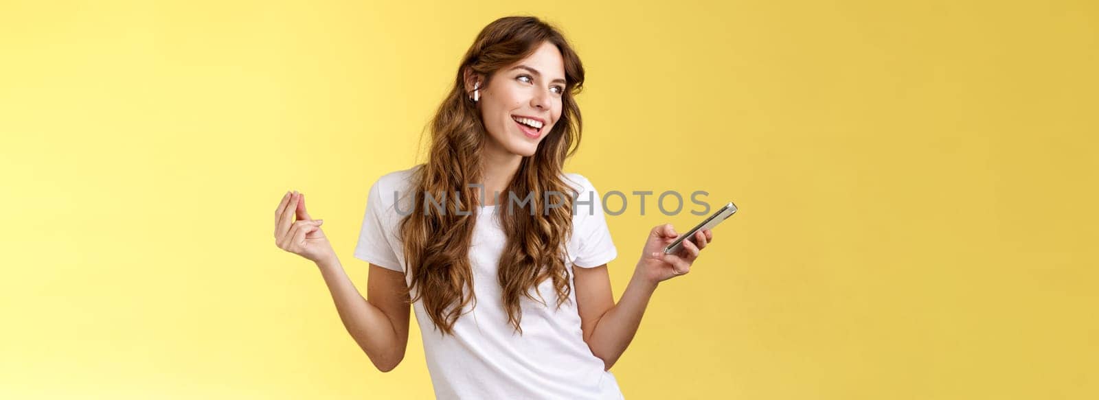 Girl enjoying music listening song wireless earbuds dancing lively carefree look away smiling broadly hold smartphone found new track partying alone soundtrack favorite movie yellow background. Lifestyle.