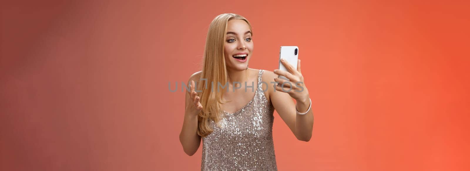 Charming elegant nice blond girl in silver dress talking video call speaking looking smartphone display amused surprised smiling happily have conversation sibling showing prom outfit by Benzoix