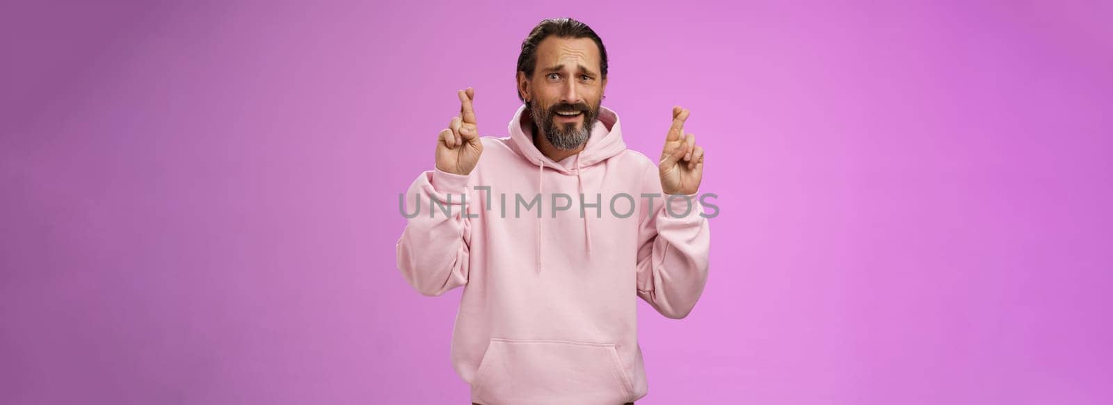 Nervous unsure hopeful handsome bearded 40s man in pink stylish hoodie cringing worried cross figers good luck make wish hope not gonna lose job supplicating praying dream come true by Benzoix