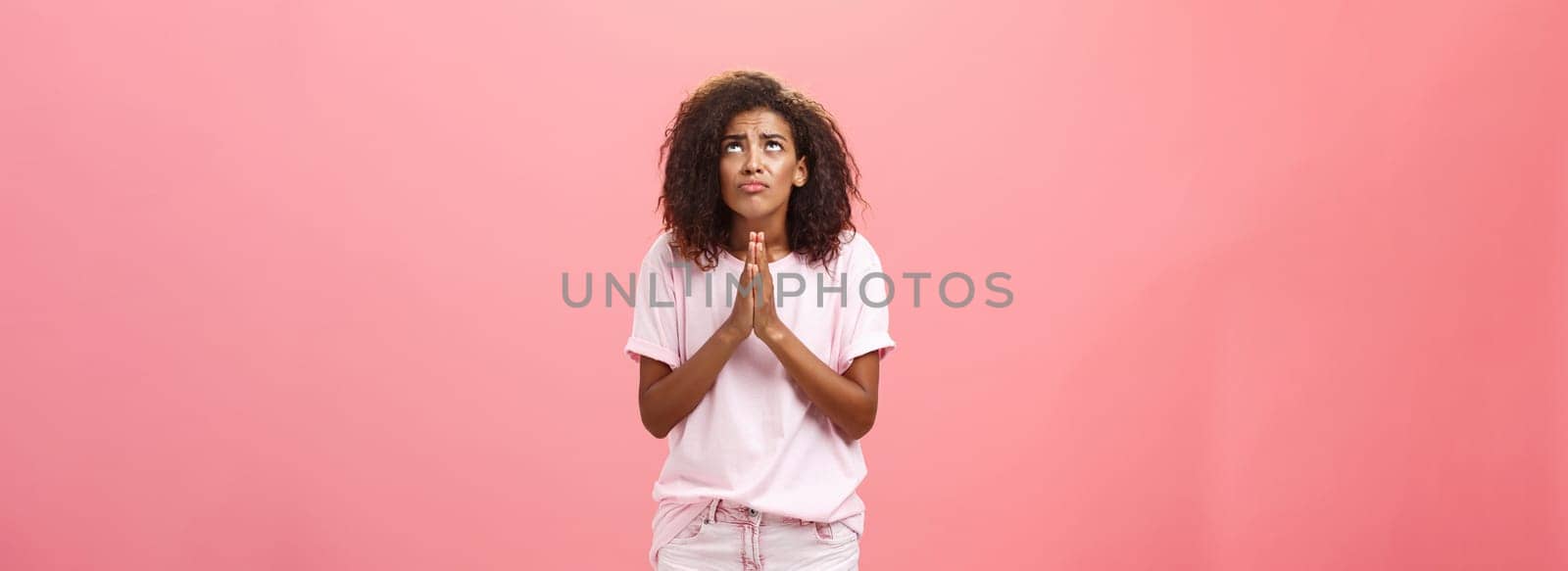 Studio shot of unhappy miserable and hopeless cute african american female holding hands in pray near chest looking up with serious-looking concerned expression making wish to god over pink wall by Benzoix