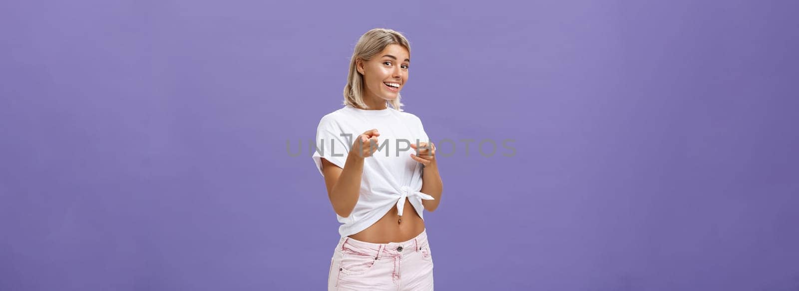 We need you in team. Joyful friendly and good-looking blonde female student in white t-shirt and shorts pointing with finger guns at camera and smiling broadly greeting friend over purple wall by Benzoix