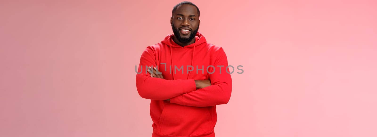 Attractive bearded african-american young guy wearing red hoodie cross arms chest smiling friendly showing friends new place grinning delighted buying own flat, standing proud pink background.