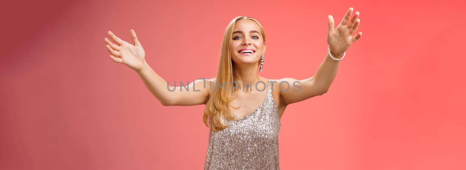 Friendly charming tender hospitable attractive birthday girl in luxury silver dress welcoming guests throw party extend arms cuddle greeting people hug give cuddle smiling broadly, red background.