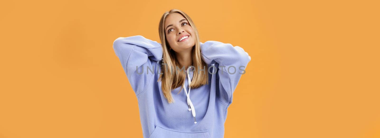 Young pleased and tender woman with tanned skin feeling cozy in warm in trendy hoodie touching back of neck smiling looking at upper right corner dreamy and delighted against orange background. Lifestyle.