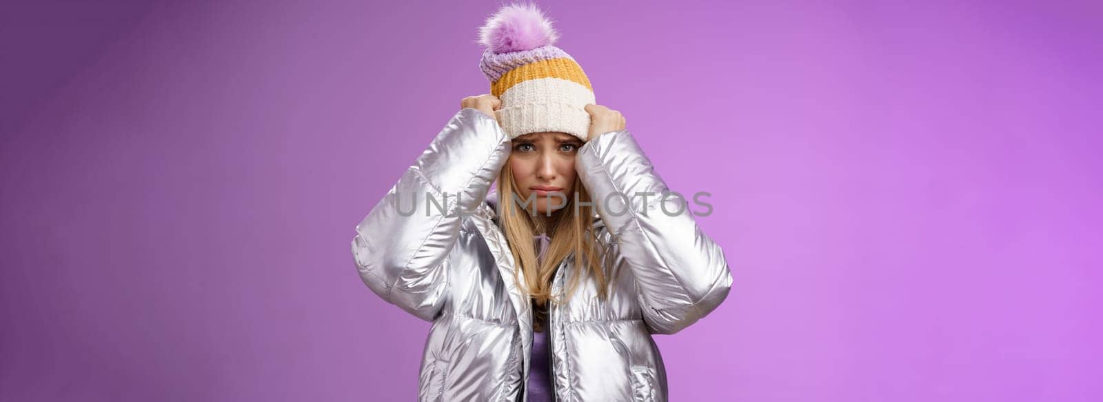 Upset gloomy insulted cute blond girl complaining offensive behaviour pulling hat forehead frowning sulking sad standing miserable pity purple background in warm stylish glittering silver jacket by Benzoix