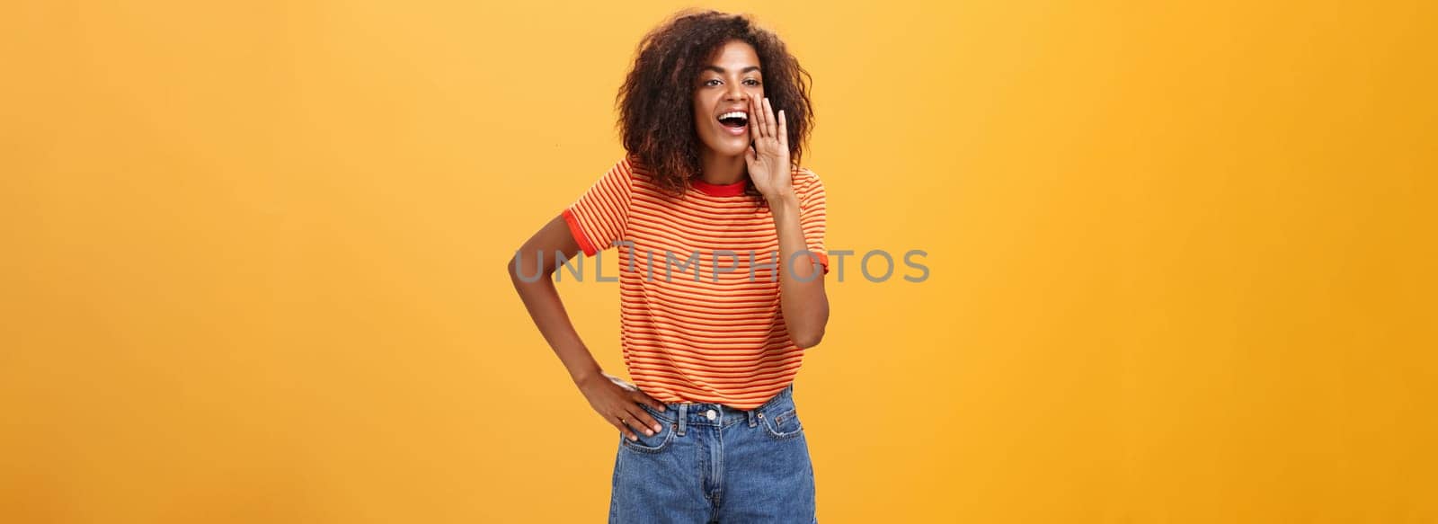 Girl calling sister come to phone yelling out loud holding palm near opened mouth to be scream louder so friend can hear holding arm on waist gazing left with carefree pleased look over orange wall by Benzoix