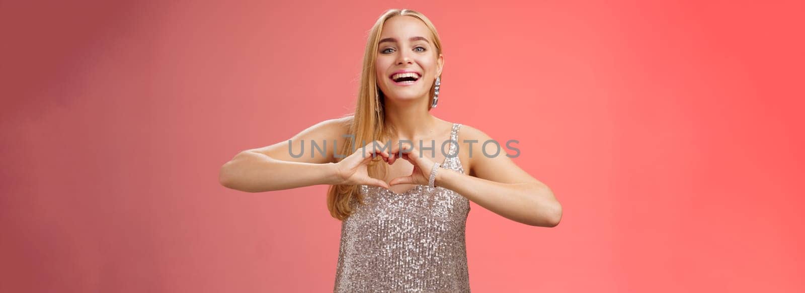 Adorable happy charming lovely blond european girl in silver dress express love positivity show heart gesture confess sympathy passionate feelings towards boyfriend smiling broadly, red background by Benzoix