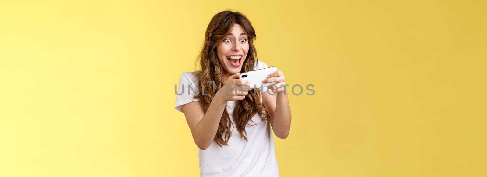 Happy upbeat playful enthusiastic curly-haired girl tempting playing stunning awesome smartphone game hold mobile phone horizontal way smiling broadly stare camera focused winning beat score by Benzoix
