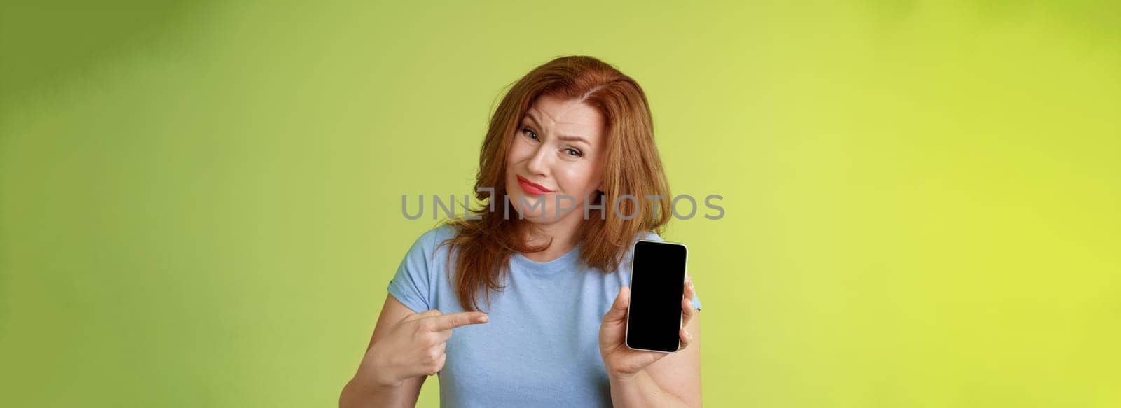 Seriously it awful. Displeased disappointed redhead mature female tilt head cringe grimacing reluctant pointing smartphone blank display index finger showing bad photograph share negative opinion.