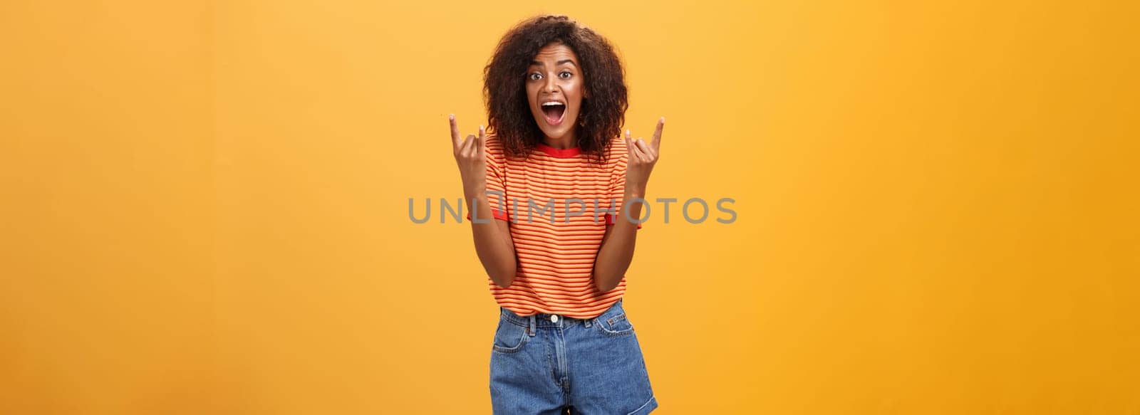 Stylish african american woman being delighted and excited before cool concert showing rock n roll gesture with both hands screaming from satisfaction and happiness cheering over orange background by Benzoix