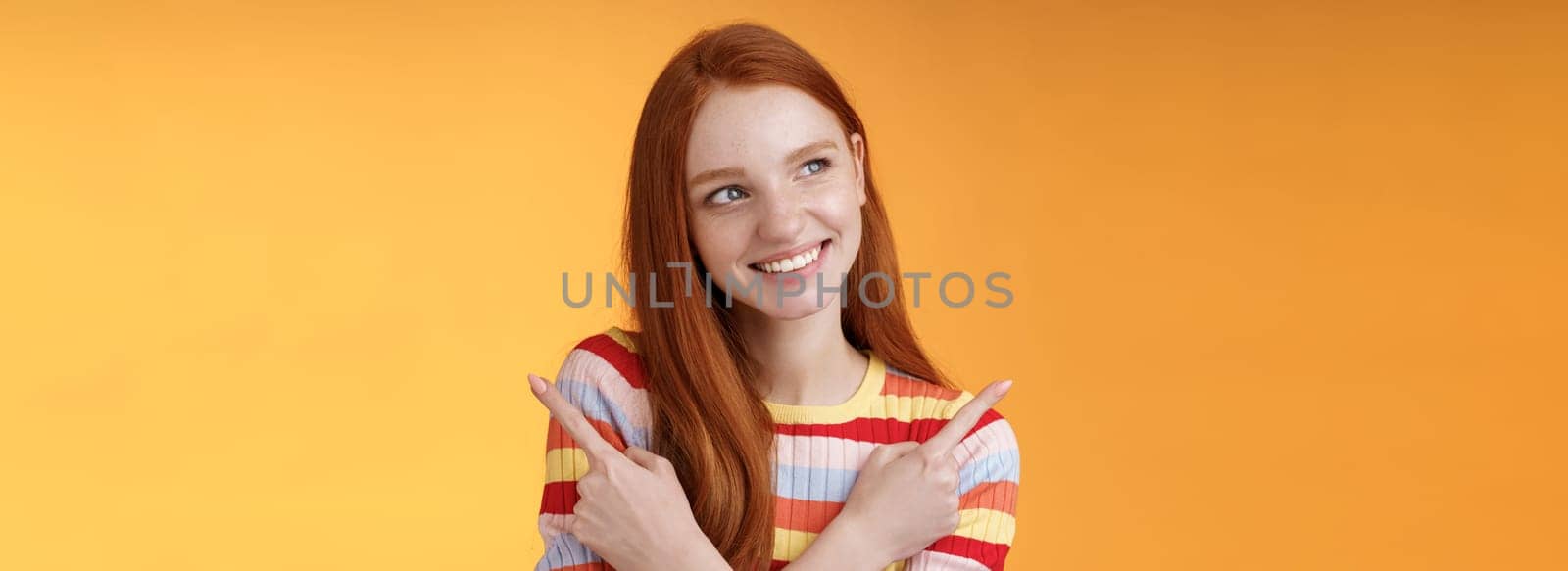 Dreamy cute redhead girl planning were go summer holidays have different choices picking variant look intrigued smiling pleased pointing sideways left right taking decision, orange background.