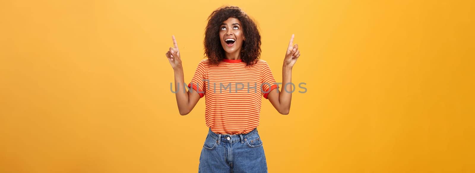 Fascinated impressed and amused good-looking charming African-American woman with afro hairstyle in trendy t-shirt and denim shorts pointing and looking up with interested look over orange wall by Benzoix