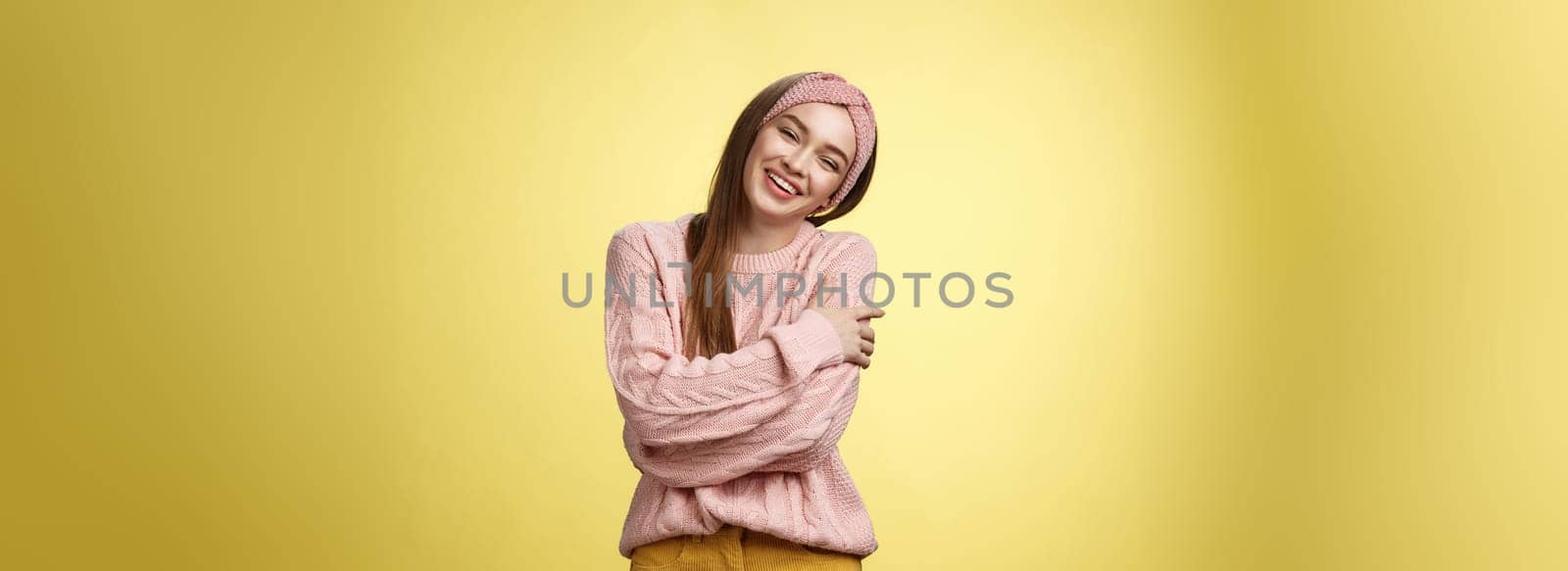 Cozy charming friendly young smiling girl in knitted warm comfortable sweater, grinning joyfully tilt head emracing herself, crossing arms, hugging feeling safe and happy, posing against yellow wall.
