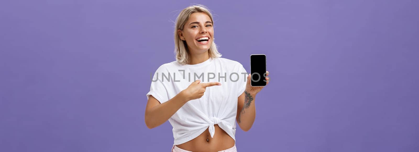 Satisfied happy beautiful tanned woman with blond hairstyle and tattoos holding smartphone showing device screen and pointing at gadget with index finger smiling at camera over purple wall. Technology and advertising concept