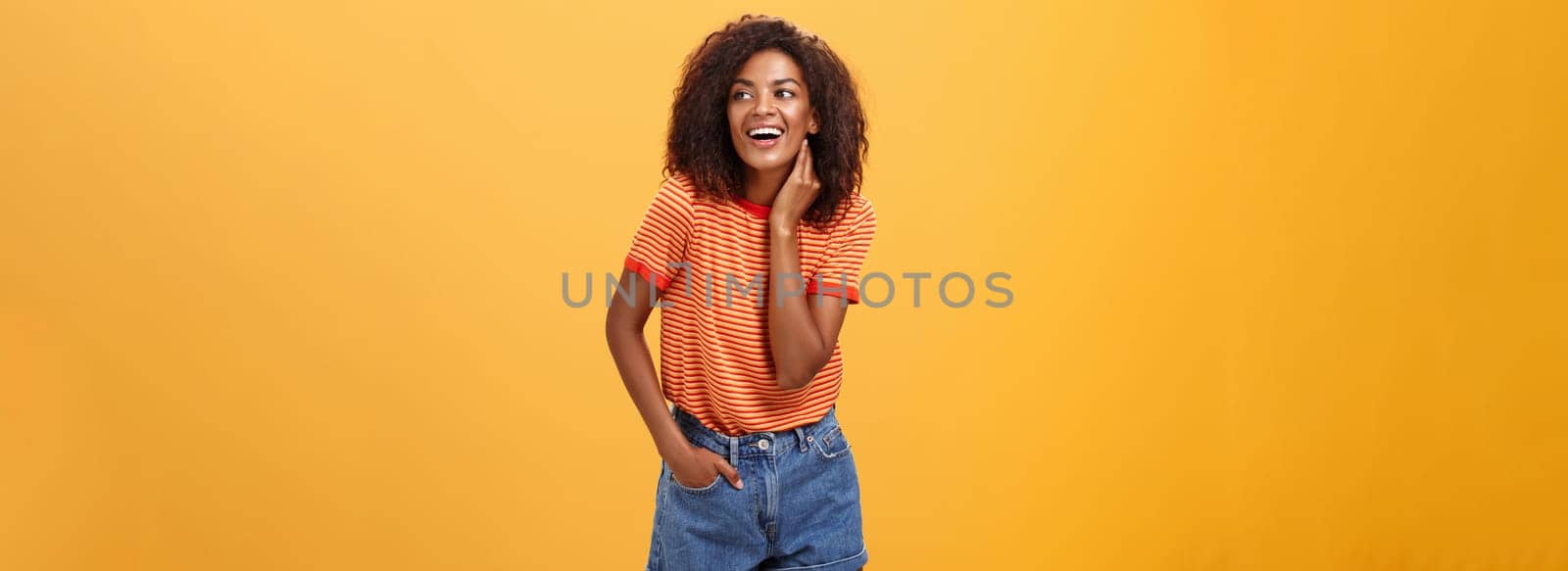 Stylish feminine and fashionable african american female model with afro hairstyle touching neck gently looking right with amused carefree expression holding hand in pocket over orange background by Benzoix