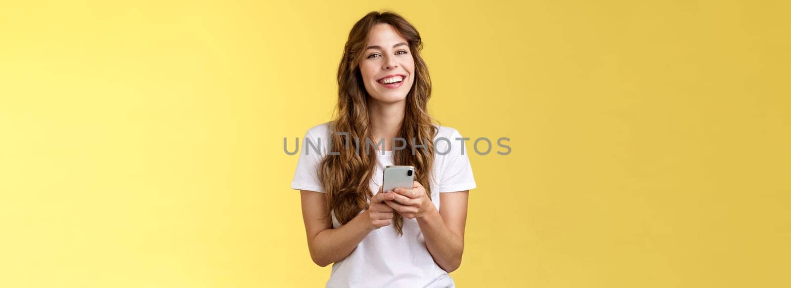 Lively enthusiastic friendly smiling happy woman using smartphone texting messaging friend checking social media feed browsing internet hold mobile phone laughing happily yellow background.