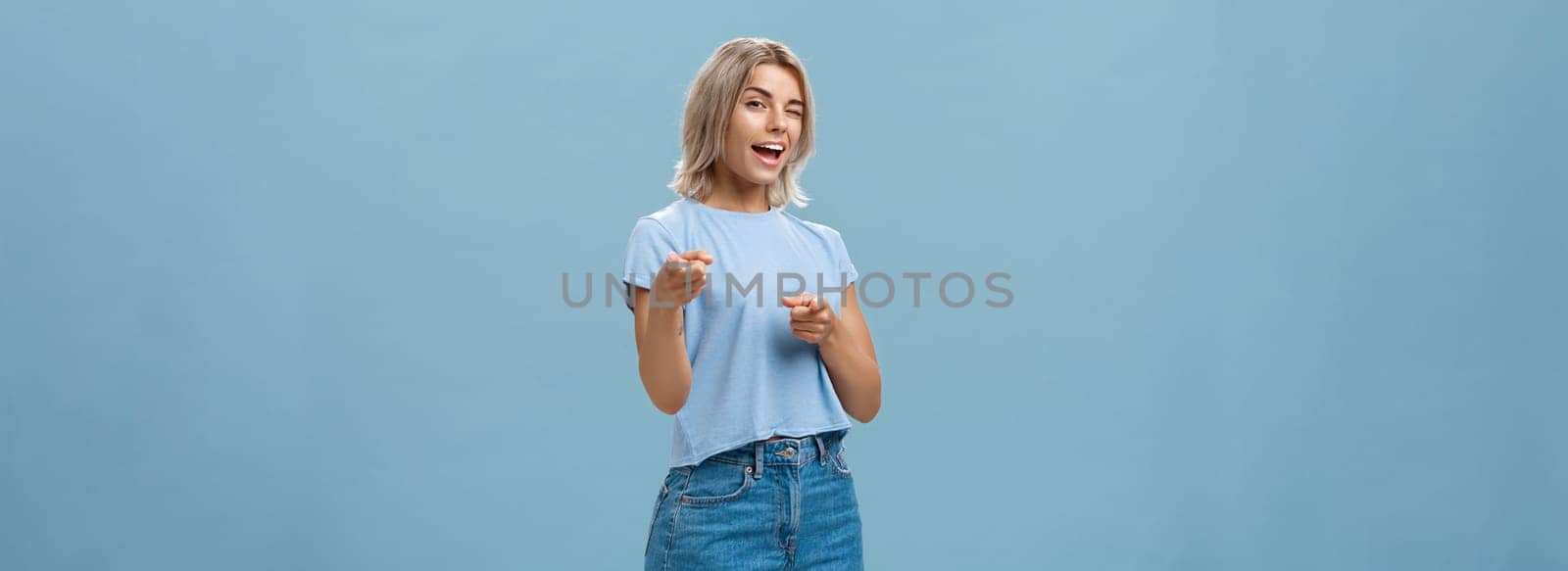 Hey I got offer for you. Portrait of attractive self-assured and playful stylish blond female with tanned skin in denim shorts and summer t-shirt winking and pointing at camera over blue background.