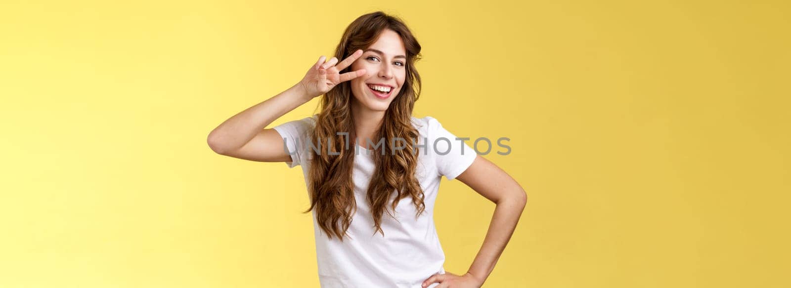 Girl having fun feel happy enthusiastic enjoy perfect summer day share positivity upbeat mood show peace victory sign near cheek tilt head smiling silly carefree stand yellow background. Lifestyle.