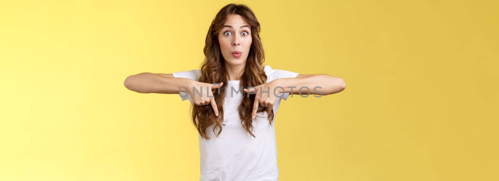 Fascinated impressed charismatic brunette female curly long haircut folding lips whistle astonished react excited thrilled telling you incredible promotion pointing down bottom yellow background.