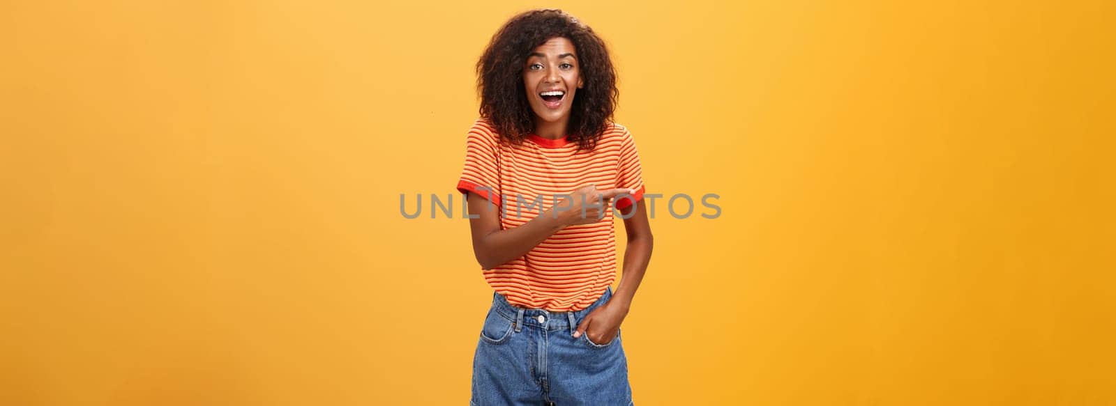 Woman laughing over funny puppy. Portrait of amused and entertained attractive stylish african-american female in casual denim shorts pointing left talking about awesome copy space over orange wall.