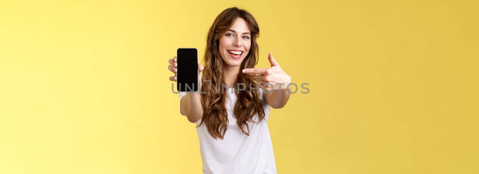Cheerful sassy good-looking sociable girl curly long hair extend arm holding smartphone pointing index finger mobile phone screen smiling broadly recommend cool app blogger promote social page by Benzoix