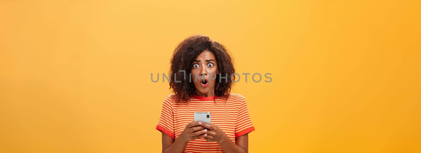 Portrait of shocked and concerned stunned dark-skinned woman with afro hairstyle popping eyes from concern and panic opening mouth holding smartphone revealing terrible information by Benzoix